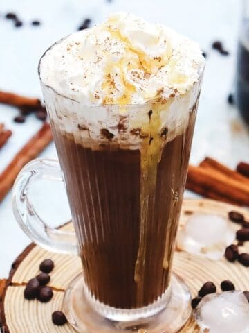 Glass mug of Copycat Starbucks Iced Cinnamon Dolce Latte topped with whipped cream and caramel, surrounded by cinnamon sticks and coffee beans.