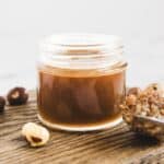 A small glass jar of brown hazelnut spread on a wooden surface with hazelnuts around it, perfect for pairing with your favorite homemade hazelnut syrup recipe.
