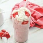 A Starbucks Copycat strawberry milkshake topped with whipped cream and fresh strawberries, served with a glass straw&mdash;just like the Strawberry Cheesecake Frappuccino recipe you love.