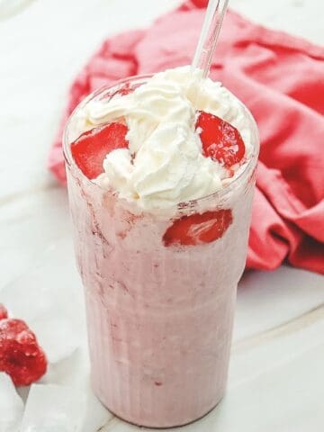 A Starbucks Copycat strawberry milkshake topped with whipped cream and fresh strawberries, served with a glass straw—just like the Strawberry Cheesecake Frappuccino recipe you love.