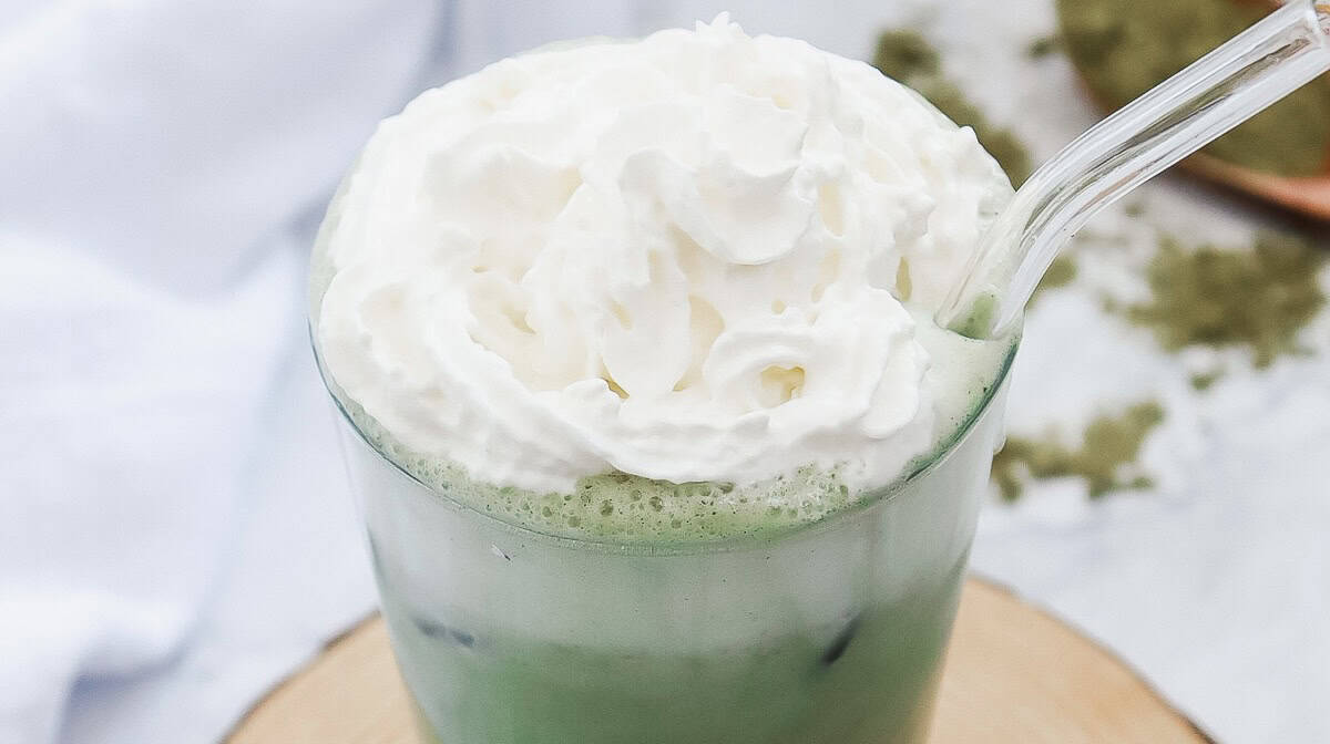 Starbucks-inspired iced matcha latte topped with whipped cream in a glass with a clear straw&mdash;a perfect copycat recipe for a Green Tea Frappuccino experience at home.