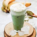 A glass of Green Tea Frappuccino with whipped cream, ice cubes, and bananas in the background sits on a wooden coaster&mdash;perfect as a Starbucks copycat recipe.