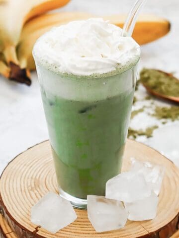 A glass of Green Tea Frappuccino with whipped cream, ice cubes, and bananas in the background sits on a wooden coaster—perfect as a Starbucks copycat recipe.