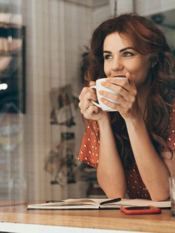 Smiling woman in a cafe holding a mug of the best ground coffee, sitting at a table with a notebook and phone.