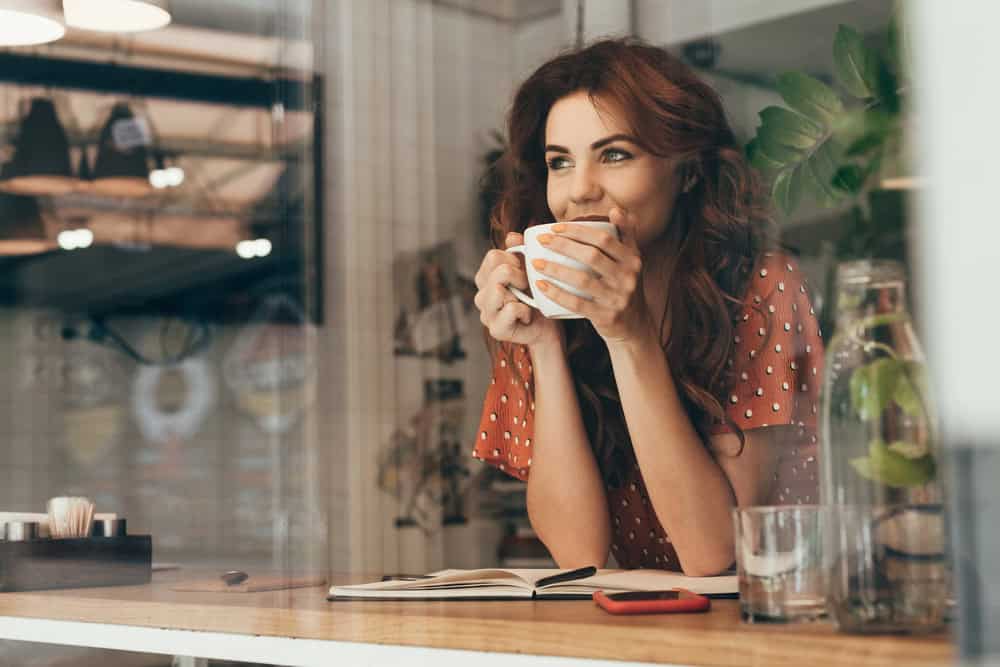 Smiling woman in a cafe holding a mug of the best ground coffee, sitting at a table with a notebook and phone.
