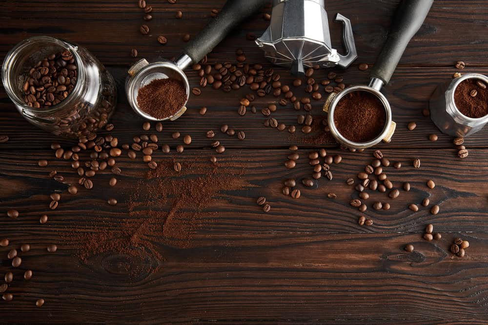 Coffee beans, best ground coffee, and moka pots on a dark wooden surface with spills and scattered beans.