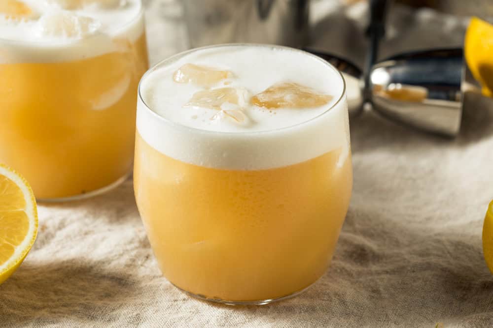 A frothy orange non-alcoholic tequila sour, served with ice cubes in a short glass, sits elegantly on a beige cloth next to fresh lemon slices.