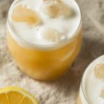A frothy whiskey sour cocktail with ice, next to a lemon half on a beige cloth surface—perfect inspiration for those who enjoy a non-alcoholic tequila sour with the same refreshing citrus twist.