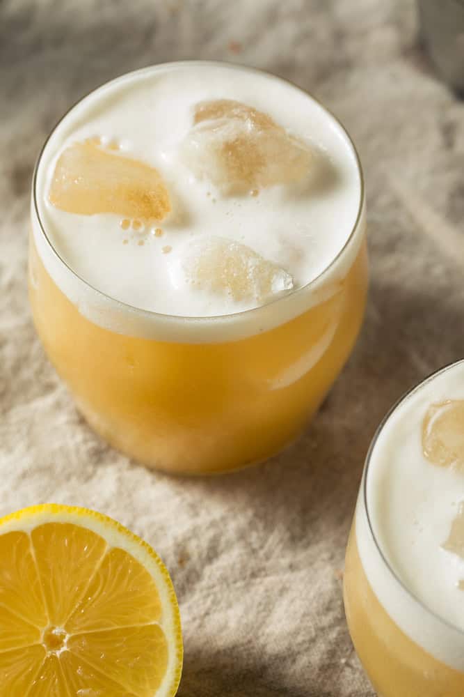 A frothy whiskey sour cocktail with ice, next to a lemon half on a beige cloth surface—perfect inspiration for those who enjoy a non-alcoholic tequila sour with the same refreshing citrus twist.
