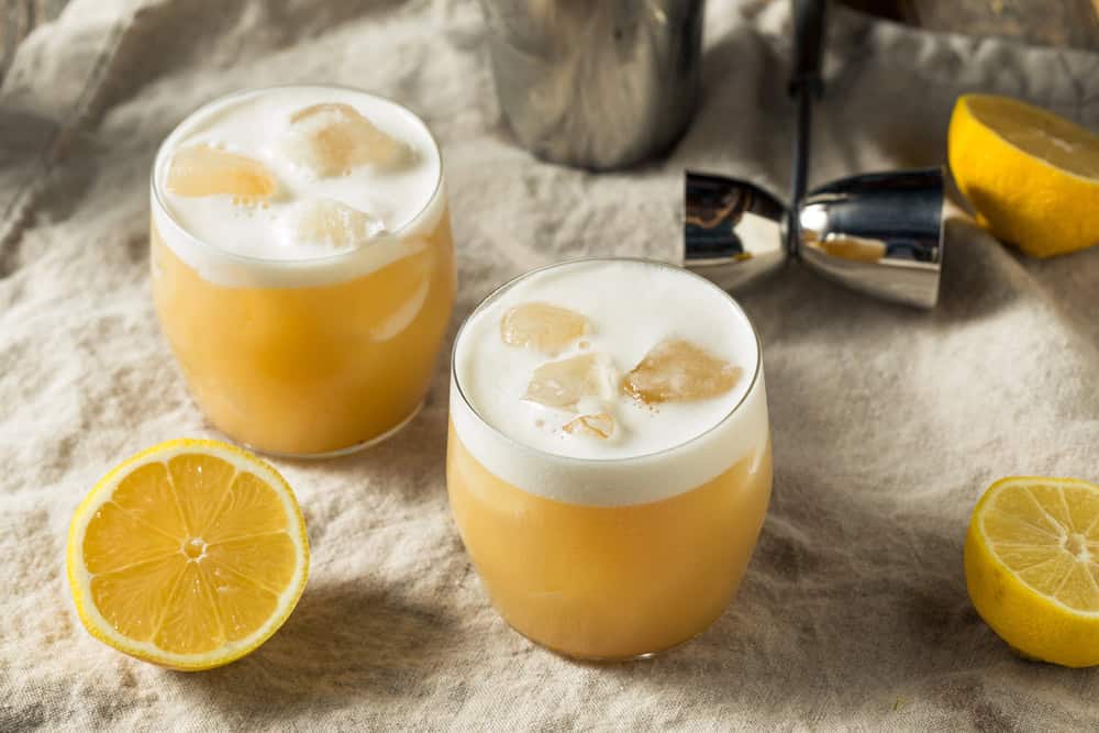 Two glasses of non-alcoholic tequila sour with ice and foam, garnished with lemon halves on a beige cloth.