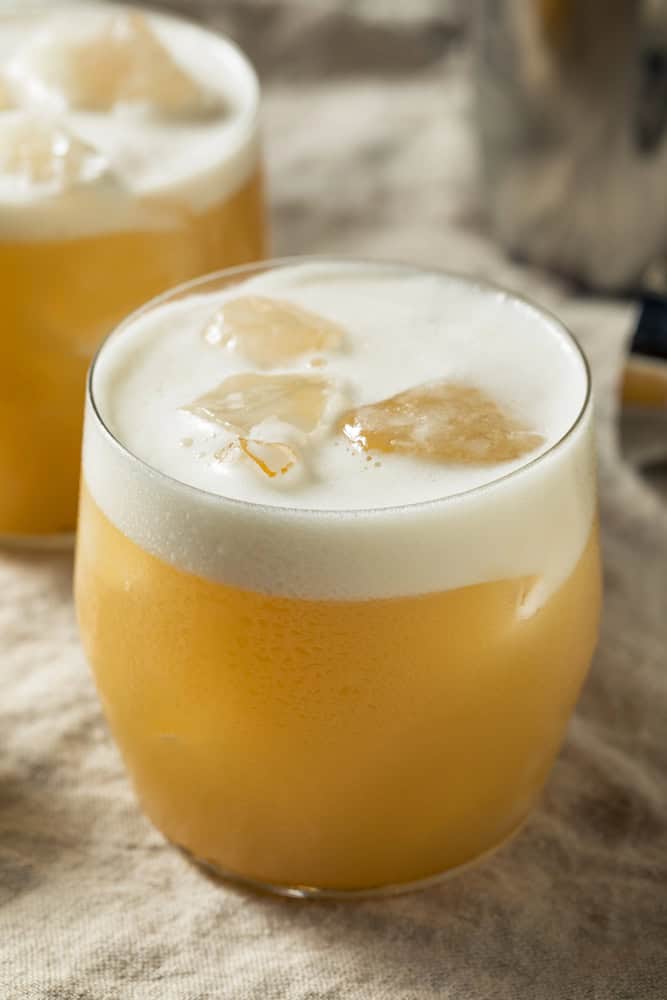 A non-alcoholic tequila sour cocktail with ice cubes and a frothy top in a round glass rests on a beige cloth surface.
