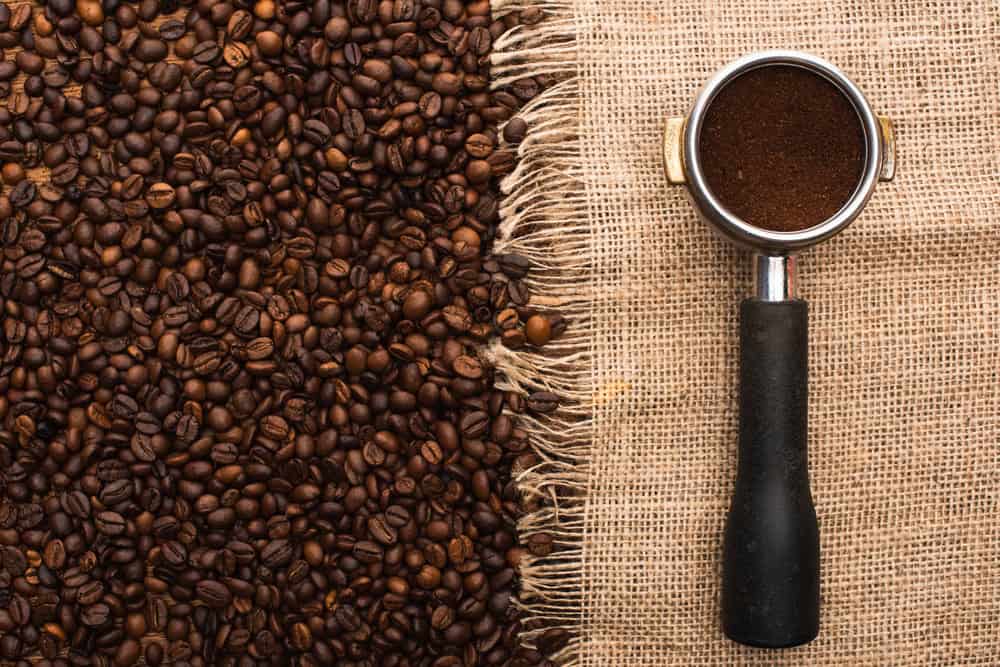 Coffee beans on burlap next to a portafilter filled with the best ground coffee, shot from above.