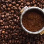 Portafilter with the best ground coffee on top of a pile of whole coffee beans, viewed from above.
