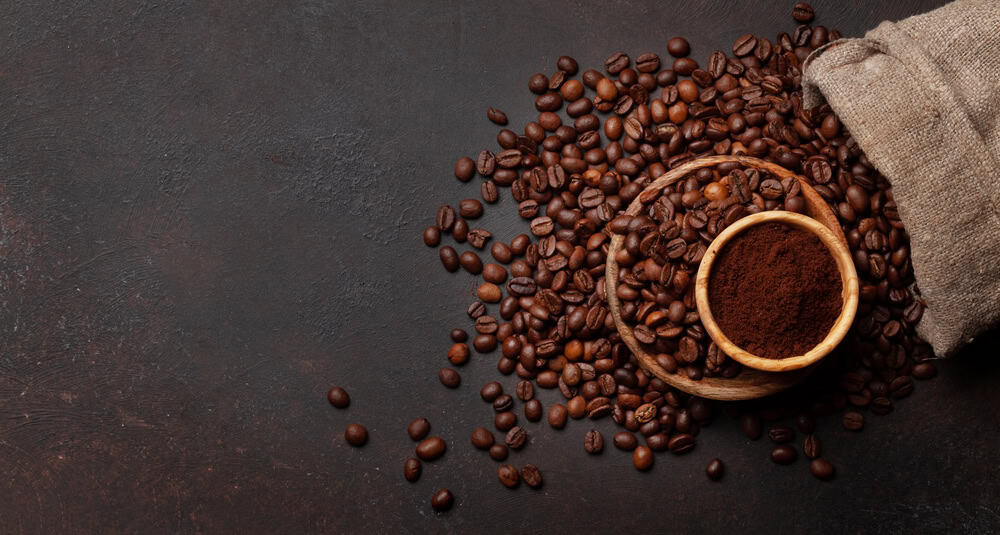 Coffee beans spilling from a burlap sack with a wooden bowl of the best ground coffee on a dark background.