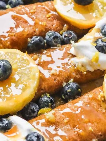 Three crepes topped with blueberries, lemon slices, whipped cream, and drizzled with syrup on a plate.