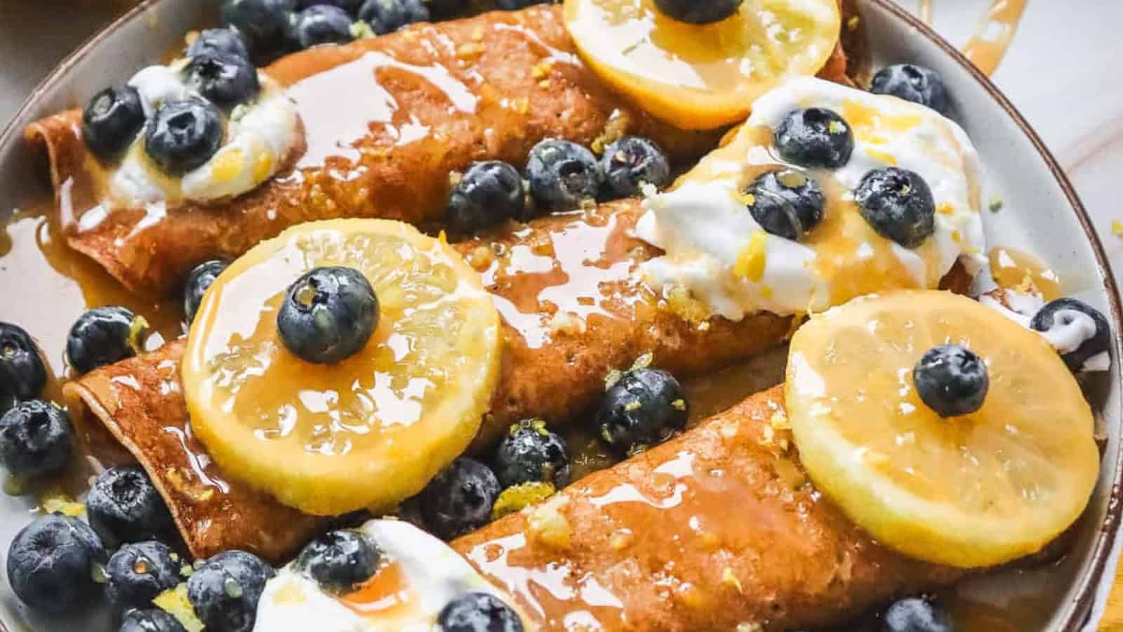 Three crepes topped with blueberries, lemon slices, whipped cream, and syrup on a plate.