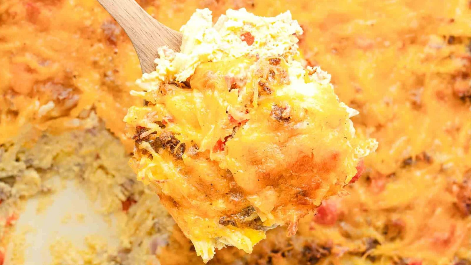A wooden spoon holds a cheesy, golden-brown casserole with visible chunks of vegetables and melted cheese.