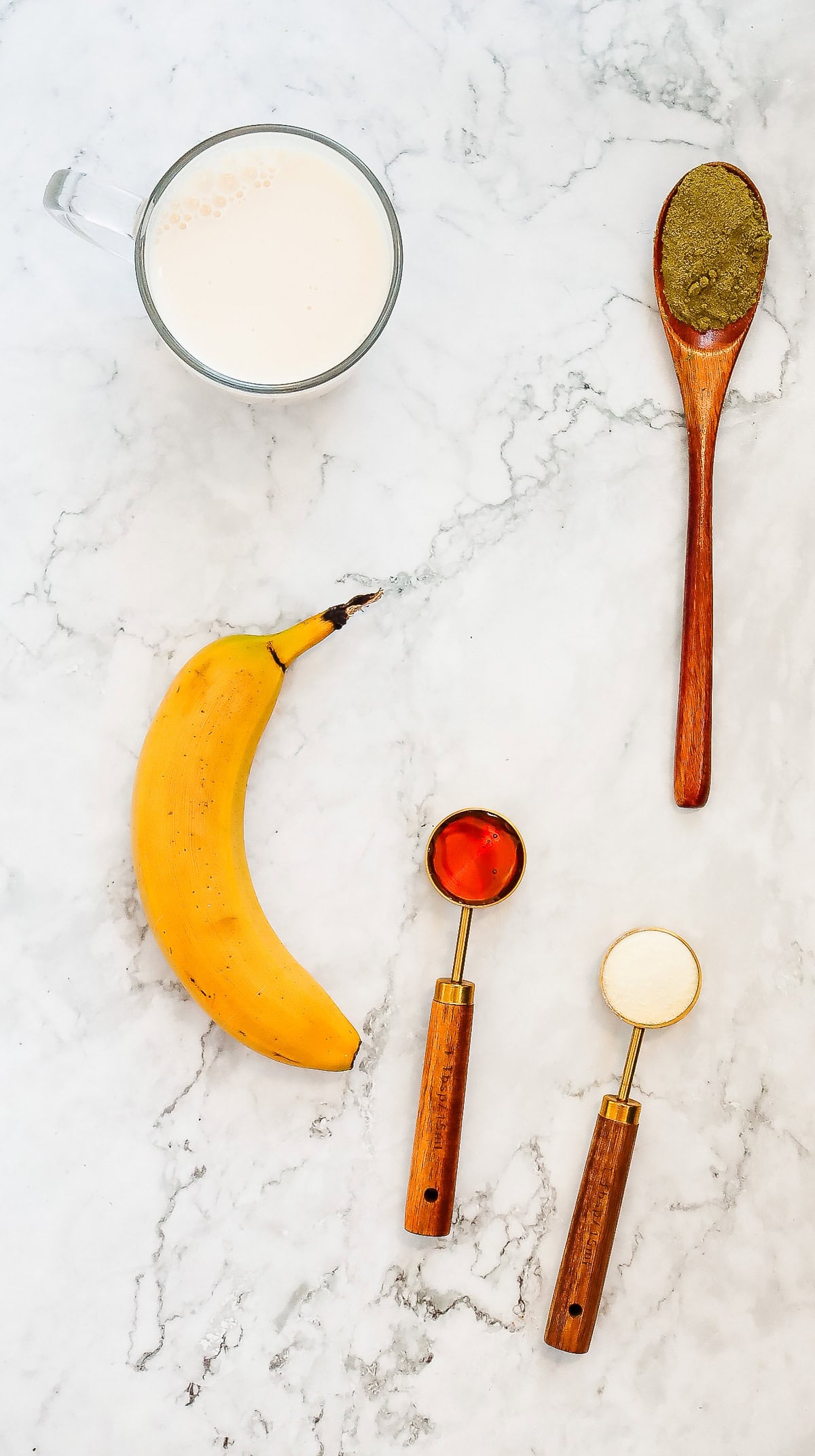 Ingredients for a Copycat Recipe: milk, a banana, protein powder, honey, and matcha powder on a marble surface, perfect for making your own Green Tea Frappuccino inspired by Starbucks.