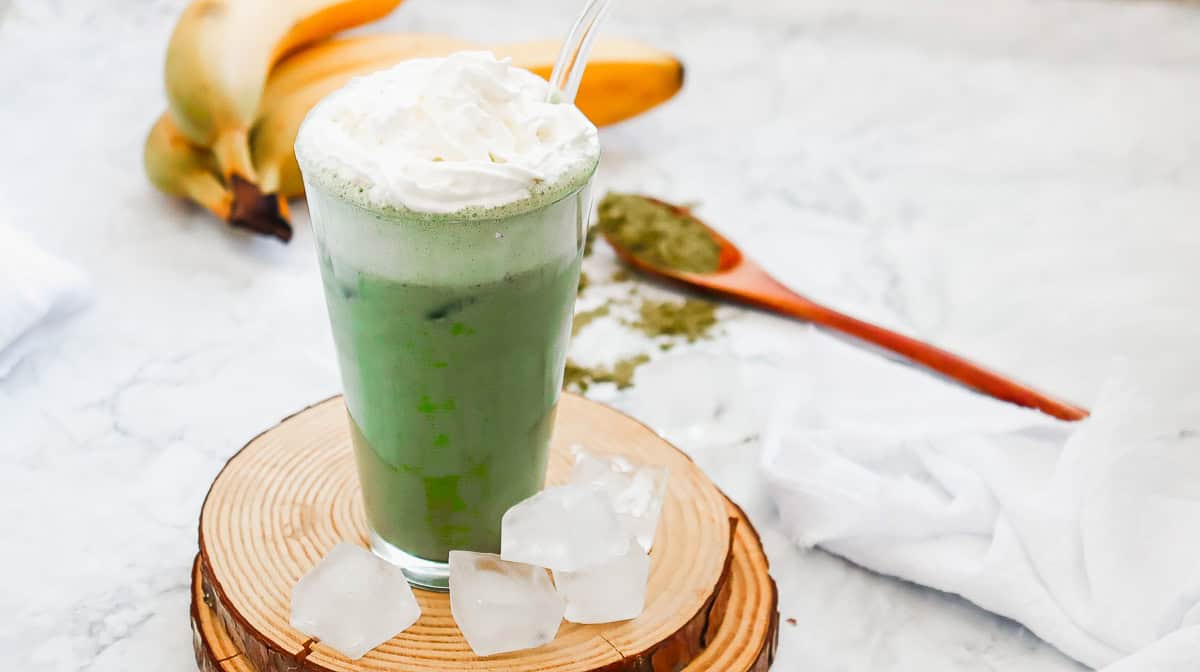 Iced matcha latte with whipped cream, ice cubes, bananas, and matcha powder on a wooden coaster&mdash;a refreshing Starbucks copycat recipe inspired by their popular Green Tea Frappuccino.