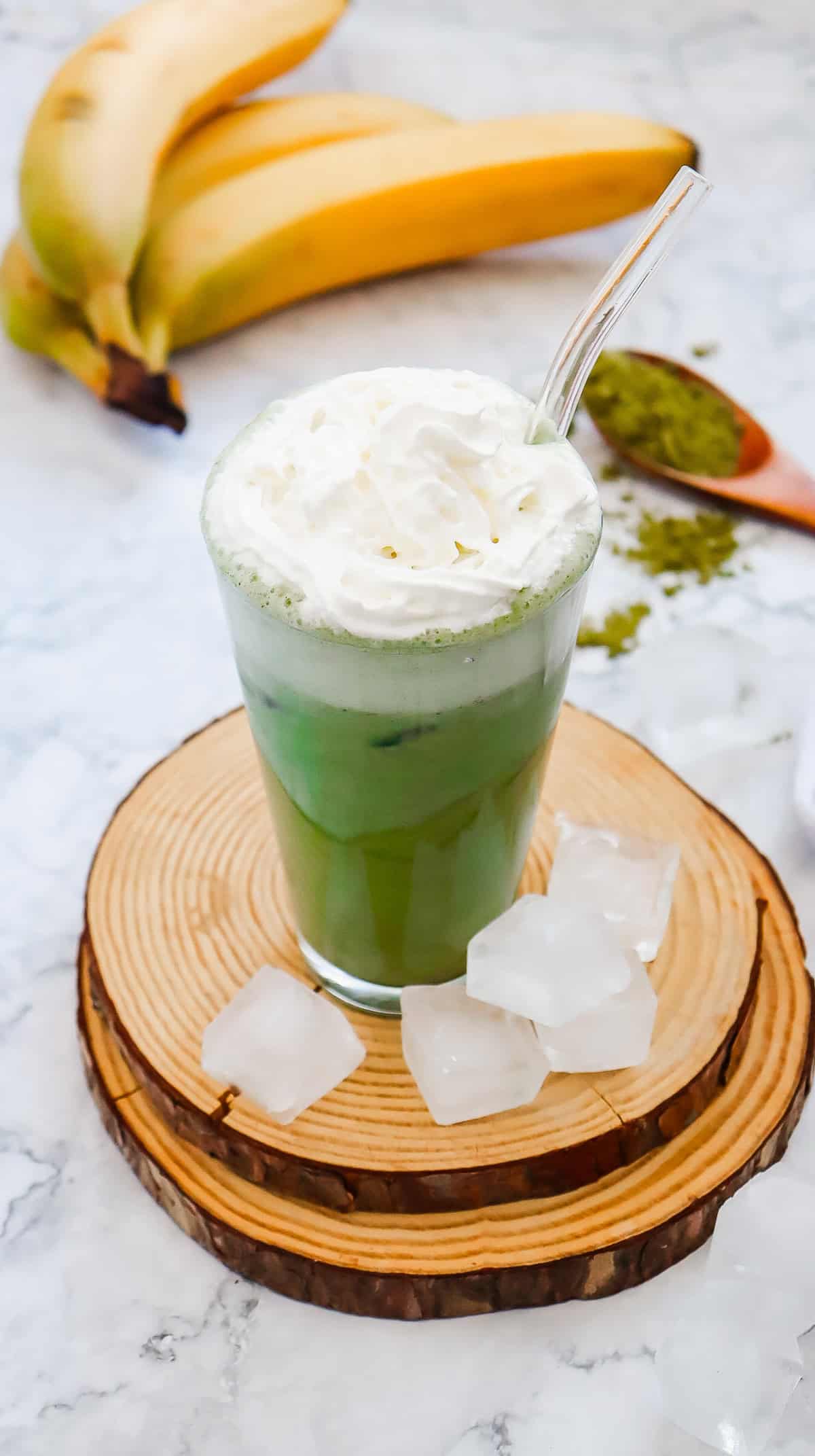 Iced matcha latte with whipped cream in a glass, surrounded by ice cubes, bananas, and matcha powder&mdash;a refreshing Starbucks Copycat perfect for crafting your own Green Tea Frappuccino recipe at home.