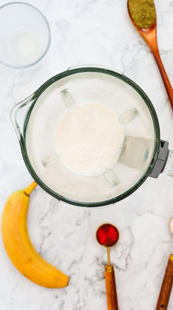 Top view of a blender with milk, surrounded by a banana, wooden spoons, a glass, and a measuring spoon&mdash;perfect setup for making a Starbucks Copycat Green Tea Frappuccino recipe.