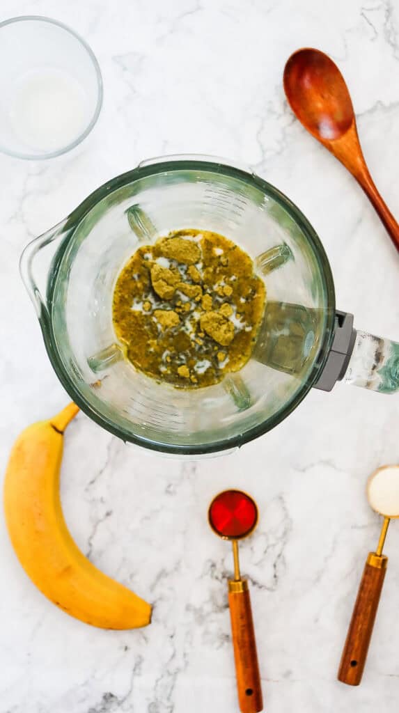A blender with green powder and milk, perfect for whipping up a Green Tea Frappuccino Starbucks copycat recipe, is surrounded by a banana, measuring spoons, a wooden spoon, and a glass.