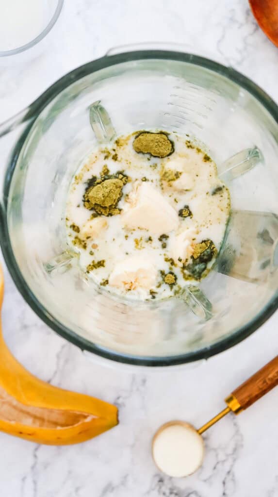 Overhead view of a blender with milk, protein powder, banana, and ice on a marble counter&mdash;perfect for creating a Copycat Recipe inspired by Starbucks' Green Tea Frappuccino, with a spoon nearby.