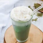 Iced matcha latte in a glass with whipped cream on top, on a wooden coaster, with bananas in the background—a delicious Starbucks Copycat recipe to enjoy at home.