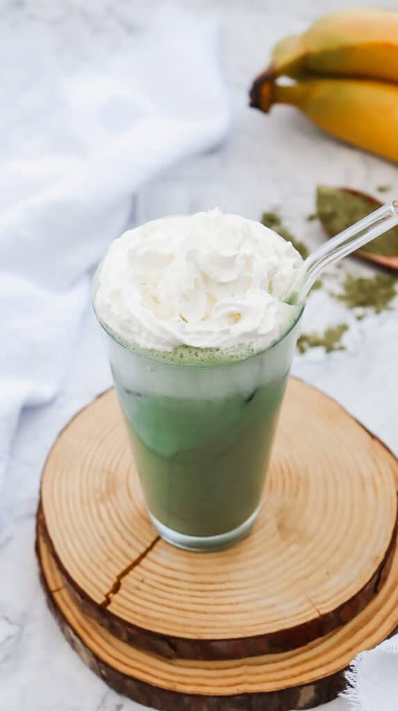 Iced matcha latte in a glass with whipped cream on top, on a wooden coaster, with bananas in the background&mdash;a delicious Starbucks Copycat recipe to enjoy at home.