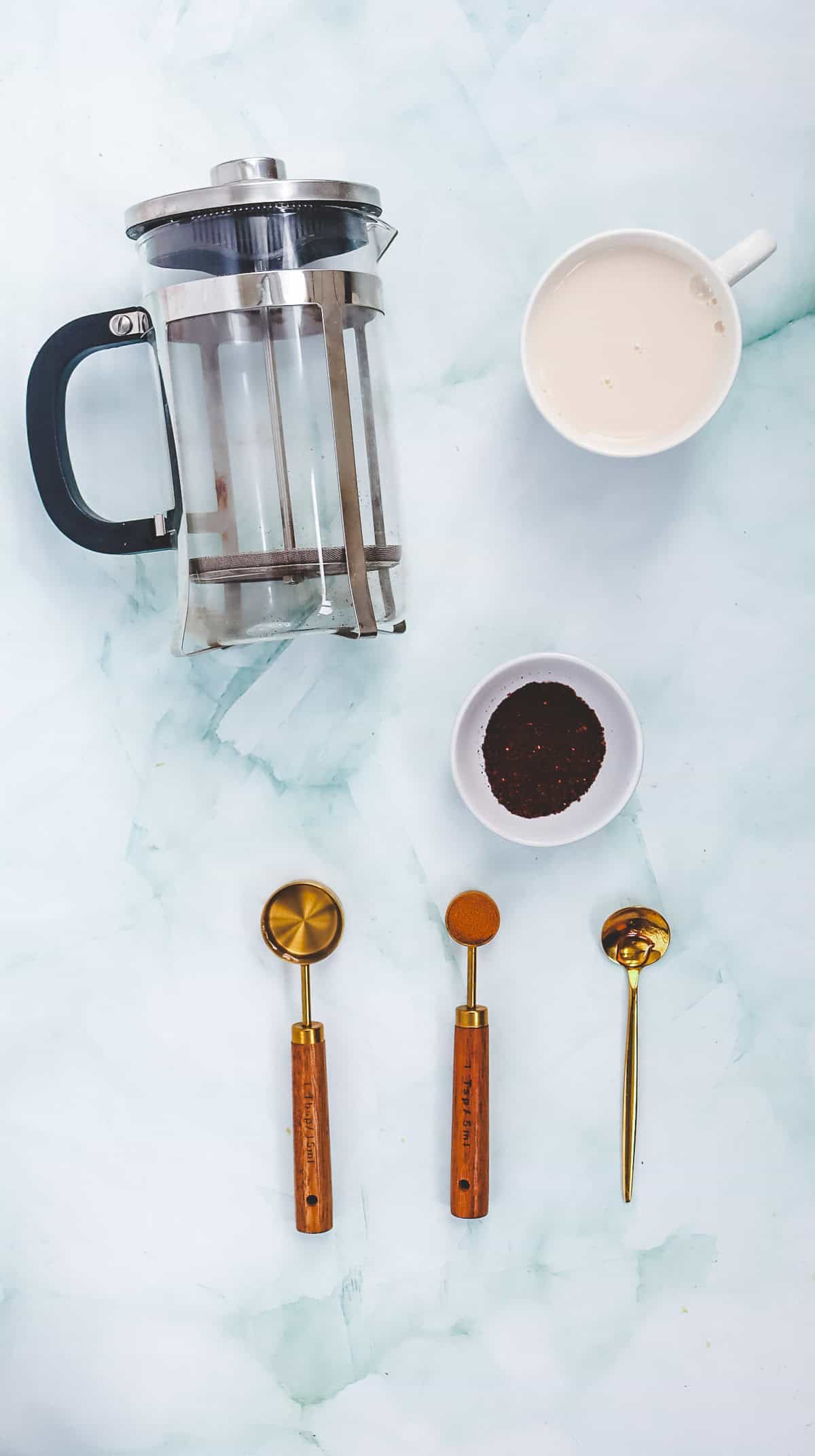 French press, cup of coffee, bowl of grounds, and three gold coffee scoops on a light marble surface&mdash;perfect for making your own Starbucks Copycat Iced Cinnamon Dolce Latte or classic latte at home.