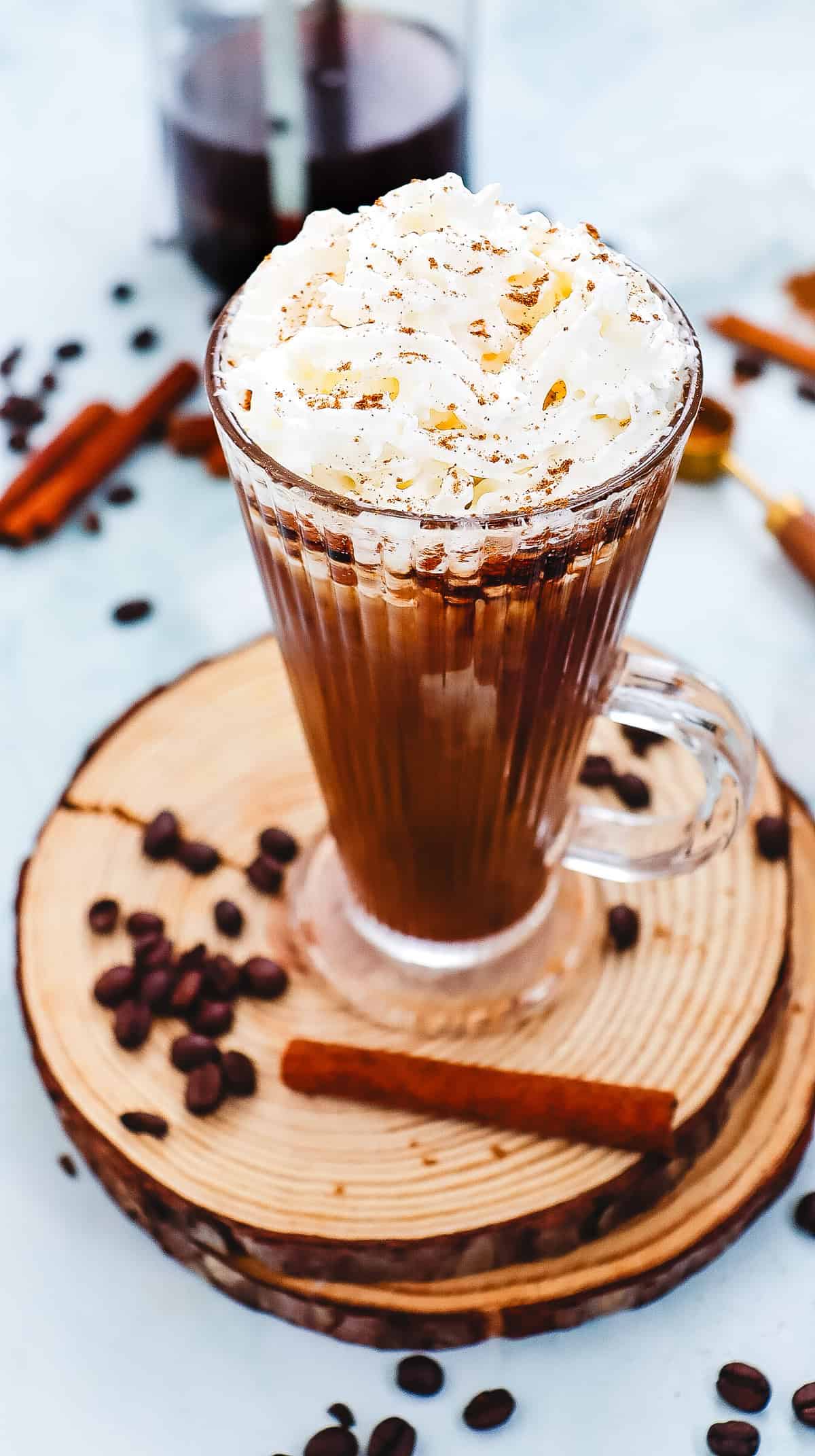 Glass mug of Cinnamon Dolce Latte topped with whipped cream, on a wooden coaster, alongside coffee beans and cinnamon sticks&mdash;a perfect Starbucks Copycat treat.