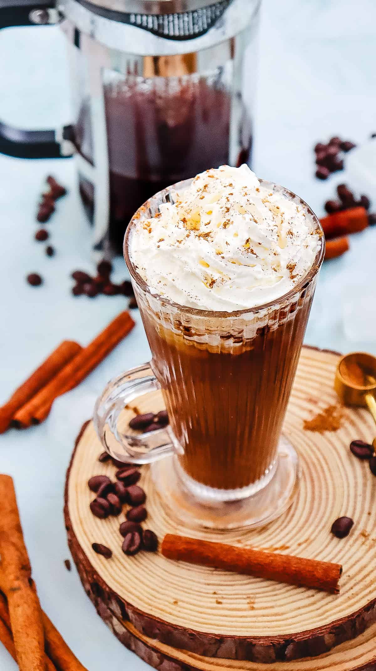 Glass mug of Iced Cinnamon Dolce Latte topped with whipped cream, surrounded by cinnamon sticks and coffee beans&mdash;a perfect Starbucks copycat treat.