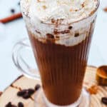 A glass mug of coffee topped with whipped cream and sprinkled spices, surrounded by coffee beans—just like a Starbucks Iced Cinnamon Dolce Latte Copycat.