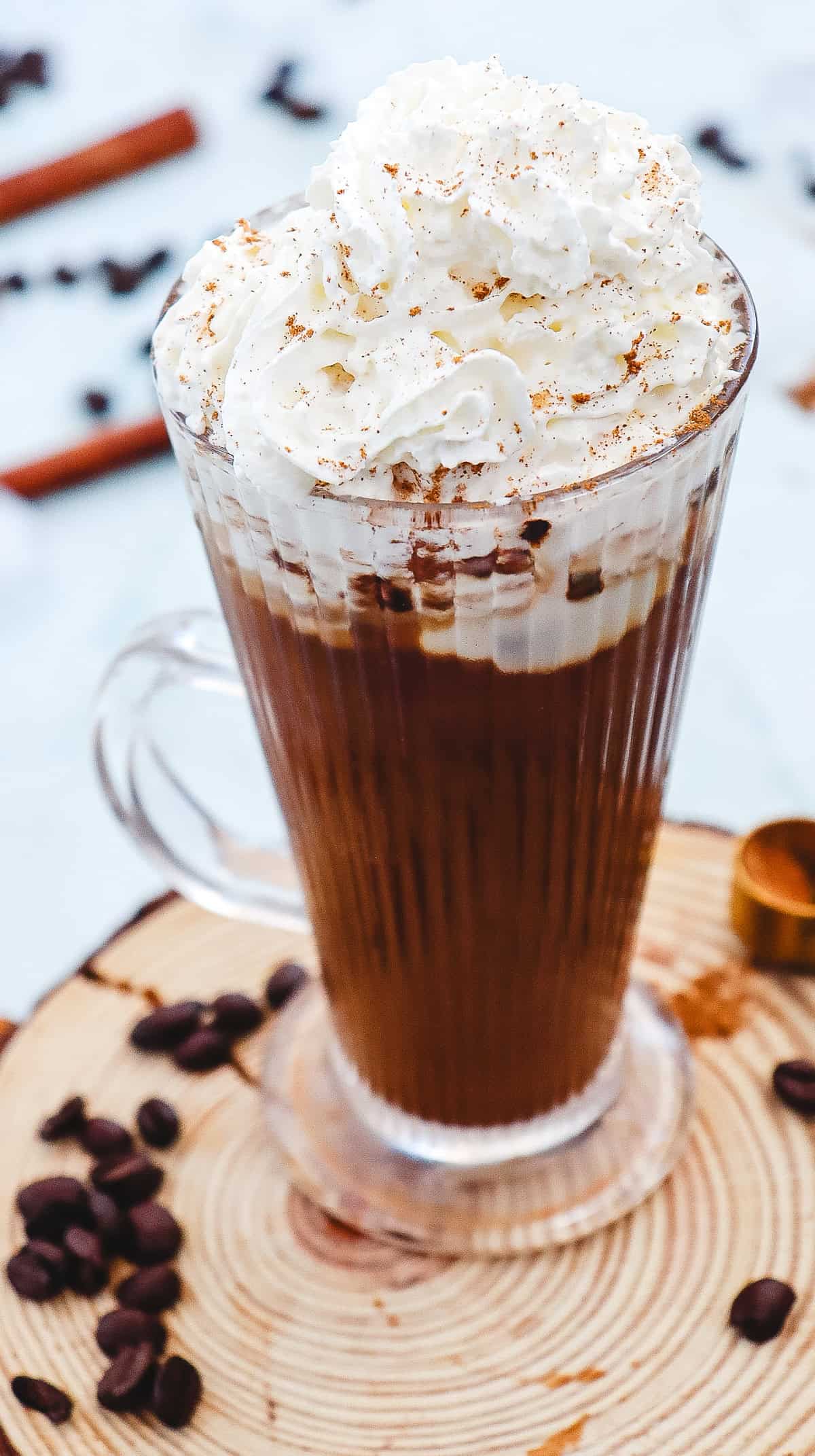 A glass mug of coffee topped with whipped cream and sprinkled spices, surrounded by coffee beans&mdash;just like a Starbucks Iced Cinnamon Dolce Latte Copycat.