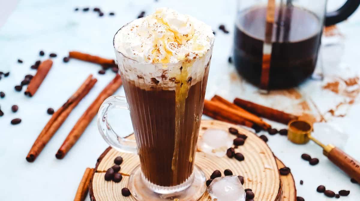 A glass mug of Iced Cinnamon Dolce Latte topped with whipped cream, surrounded by cinnamon sticks and coffee beans&mdash;a perfect Starbucks copycat treat.