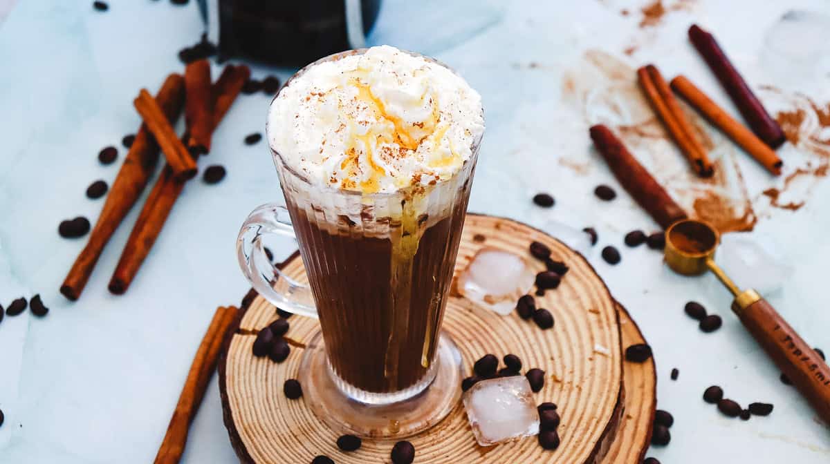 A glass mug of Cinnamon Dolce Latte topped with whipped cream and cinnamon, surrounded by coffee beans and cinnamon sticks&mdash;a perfect Starbucks Copycat treat.