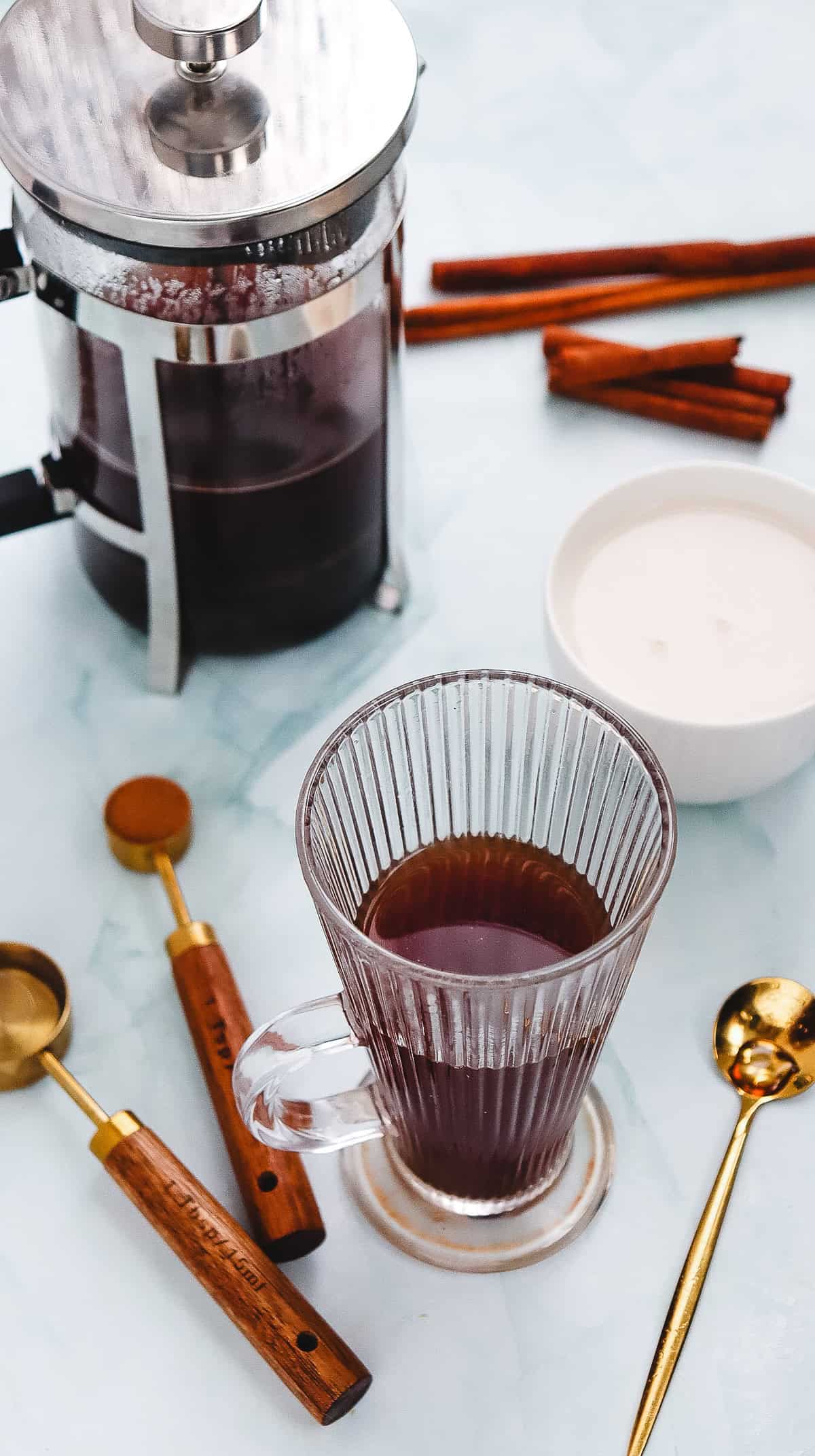 A glass mug of black coffee beside a French press, cinnamon sticks, a bowl of cream, and measuring spoons&mdash;perfect for creating your own Starbucks Copycat Cinnamon Dolce Latte at home.