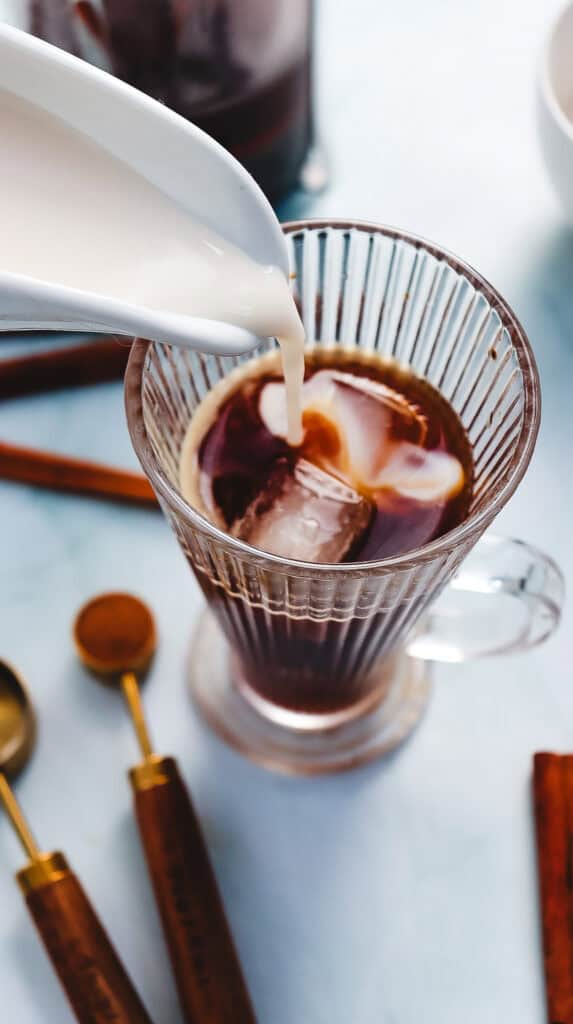 Milk is being poured into a glass of Iced Cinnamon Dolce Latte with measuring spoons and cinnamon sticks nearby, creating a delicious Starbucks Copycat treat.