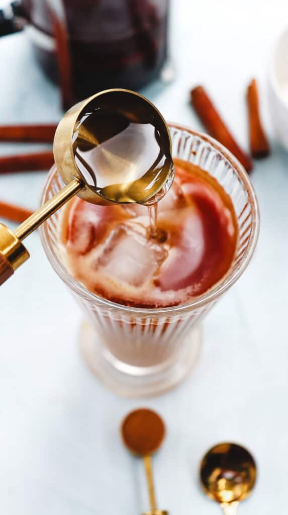 A spoon pours syrup into an iced coffee in a glass, with cinnamon sticks in the background&mdash;perfect for making your own Iced Cinnamon Dolce Latte, a delicious Starbucks Copycat treat.