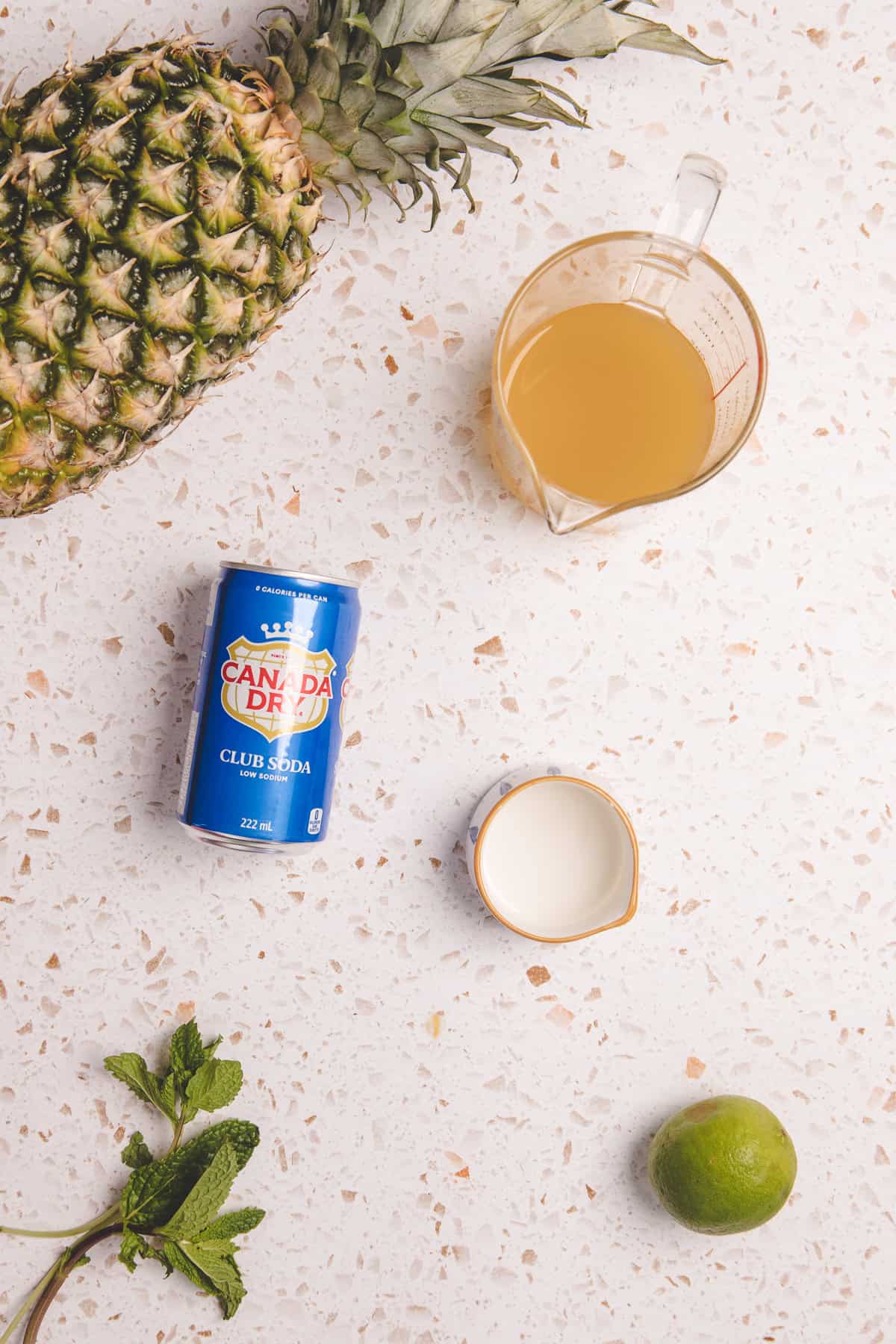 Ingredients on a counter: pineapple, club soda, pineapple juice, cream, lime, and mint leaves—everything you need for a refreshing Pineapple Mocktail or mocktail recipe.