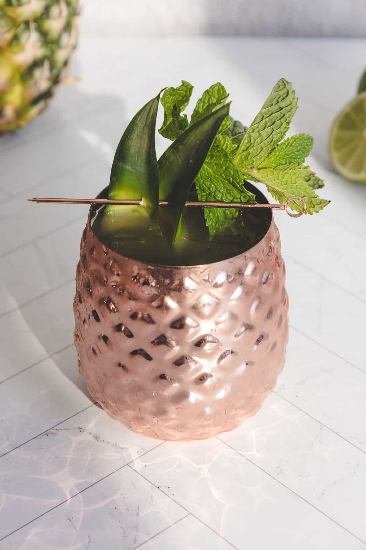 A copper pineapple cup holds a vibrant green mocktail, topped with mint and garnish, displayed on a white surface. Perfect for trying a refreshing new recipe.