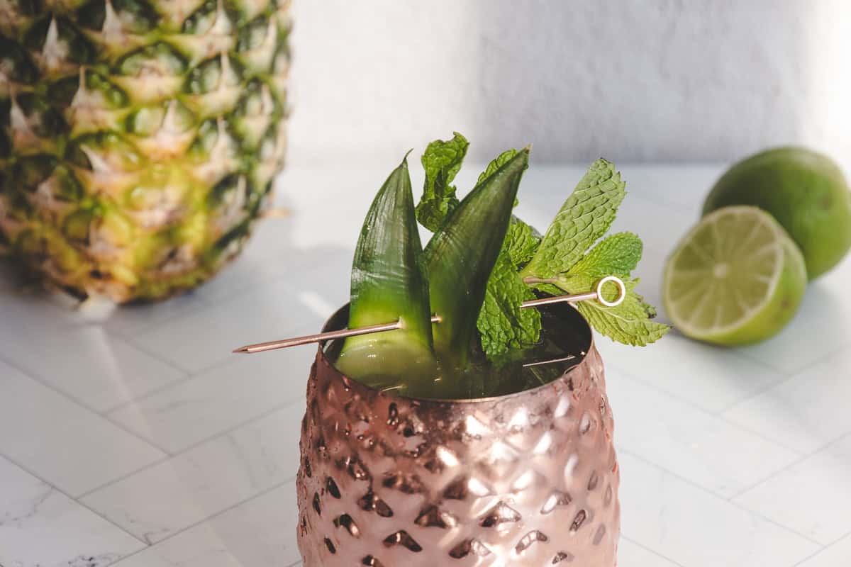 A copper cup filled with a refreshing pineapple mocktail, garnished with pineapple leaves and mint, sits on a white surface.
