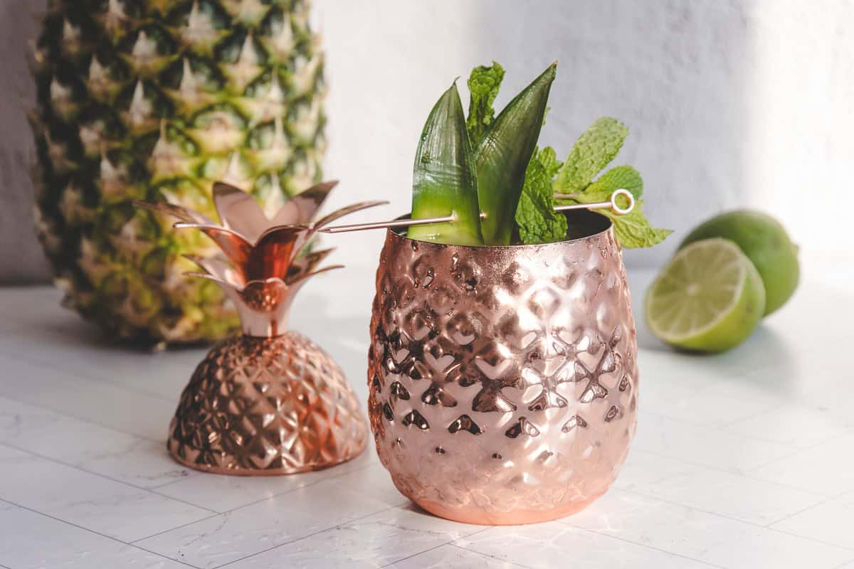 Copper pineapple cup with a lid, filled with a Pineapple Mocktail Recipe—mint and pineapple leaves garnish; fresh pineapple and lime in the background.