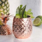 Copper pineapple cup with leafy garnish, next to its lid, a pineapple, and lime halves on a white surface—perfect for serving your favorite Pineapple Mocktail.