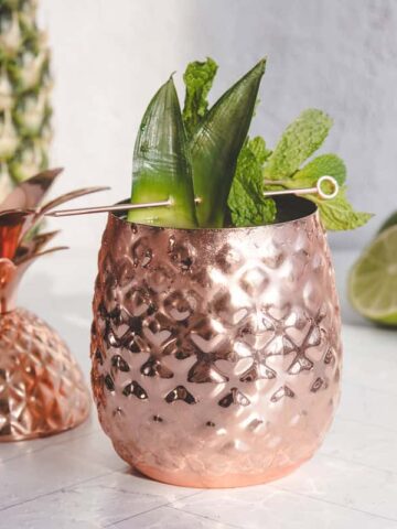 Copper pineapple cup with leafy garnish, next to its lid, a pineapple, and lime halves on a white surface—perfect for serving your favorite Pineapple Mocktail.