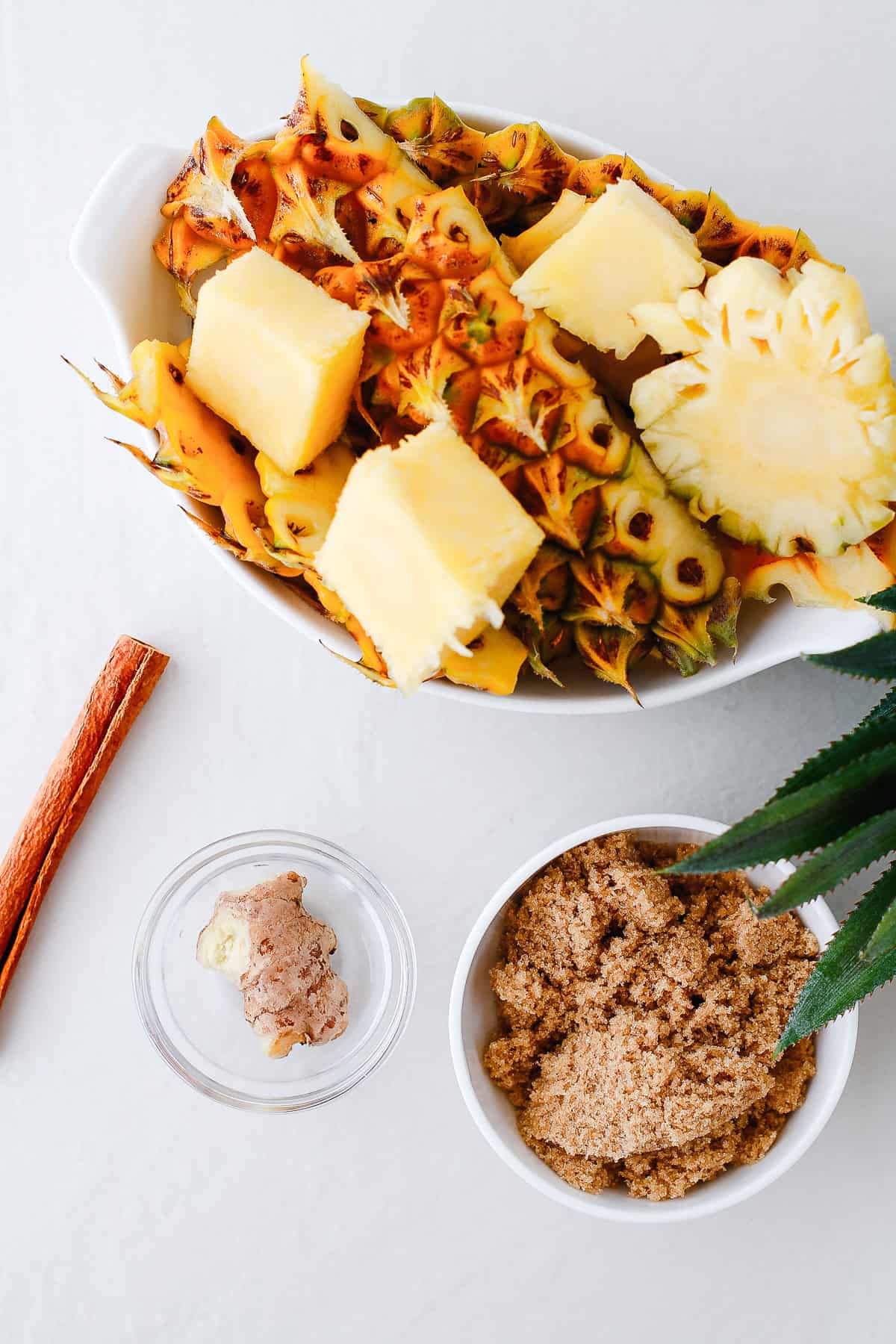 Bowl of pineapple chunks and peel, brown sugar, ginger, and a cinnamon stick on a white surface—perfect ingredients for trying an authentic tepache recipe or making your own refreshing fermented drink.