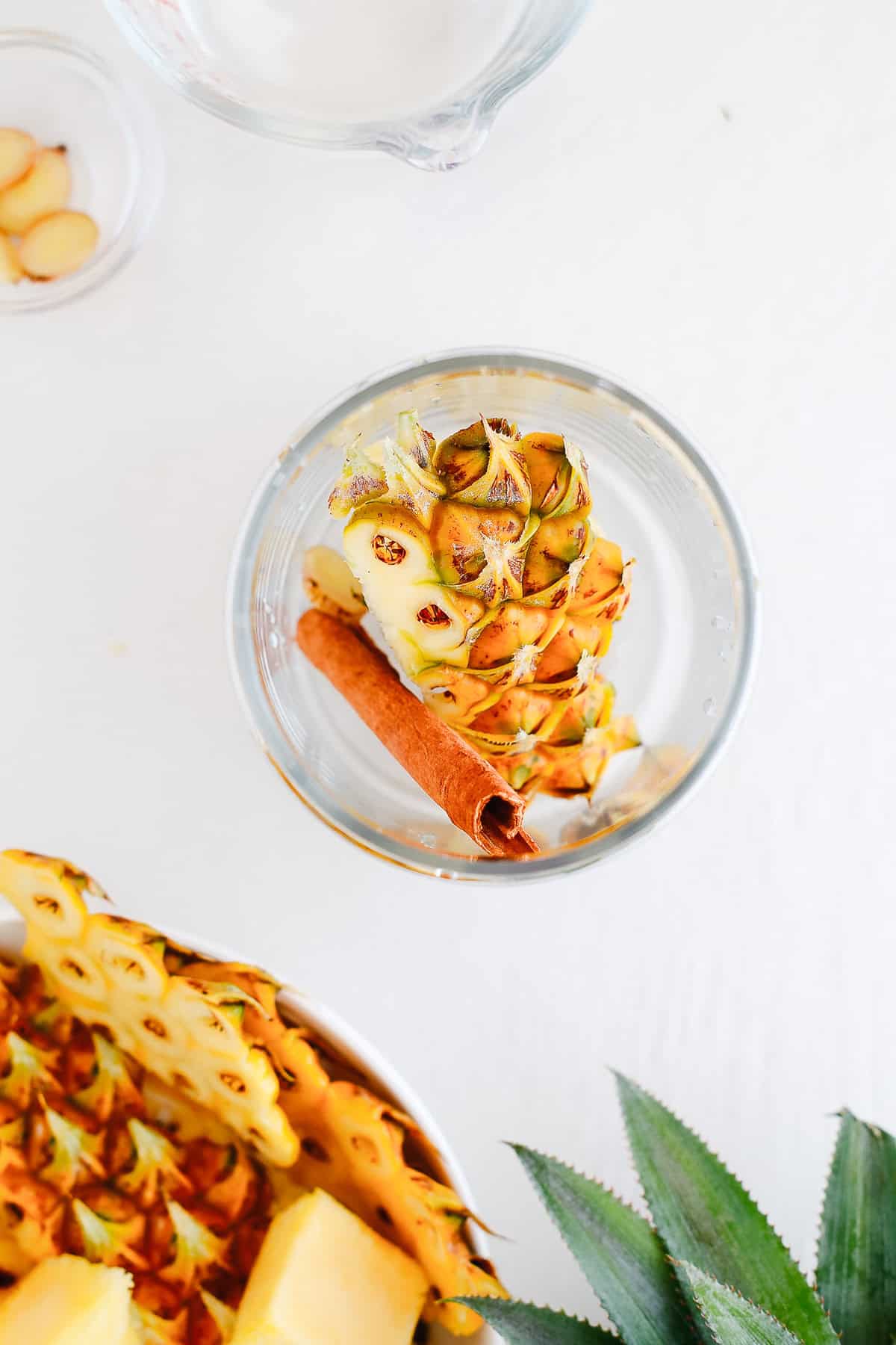 A glass of water with pineapple rind and a cinnamon stick, next to fresh pineapple pieces on a white surface—perfect for crafting a refreshing fermented drink like tepache recipe.
