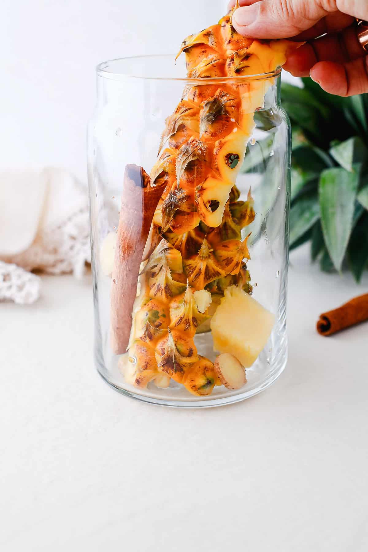 A hand places pineapple rind, a cinnamon stick, and pineapple chunks into a clear glass jar to begin this delicious tepache recipe for a refreshing fermented drink.