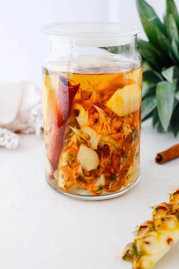 A glass jar filled with pineapple peels, cinnamon stick, and liquid—perfect for a traditional Tepache recipe—sits nearby a fresh pineapple top.
