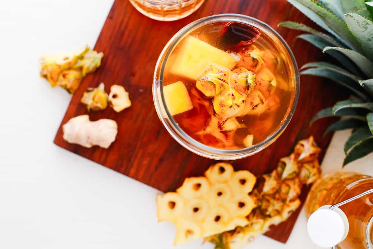 Top view of a glass of fermented pineapple drink with pineapple chunks on a wooden board, surrounded by fresh pineapple and ginger, perfect for trying your own tepache recipe.